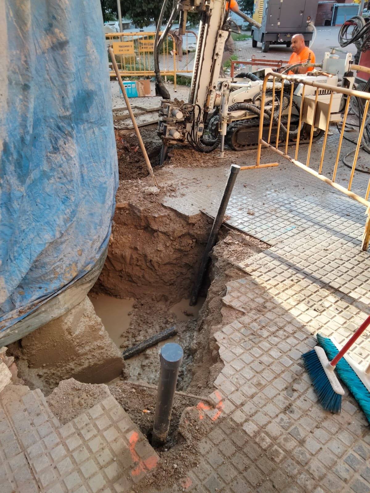 Perforadora sobre cadenas junto a la fachada de un edificio protegida con lona, con un operario a los mandos y, en primer plano, un tubo de inyección saliendo de una cata abierta en el acerado.