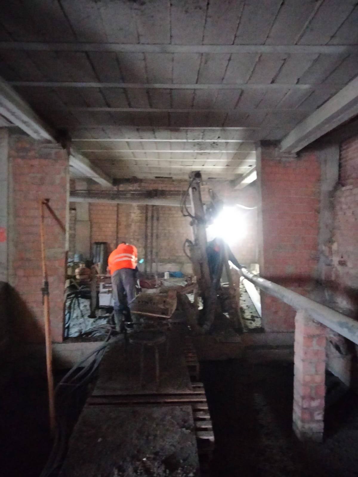 Operario con chaleco naranja junto a una perforadora iluminada por un foco, dentro de una planta baja vaciada, entre pilares de ladrillo y bajo el forjado.