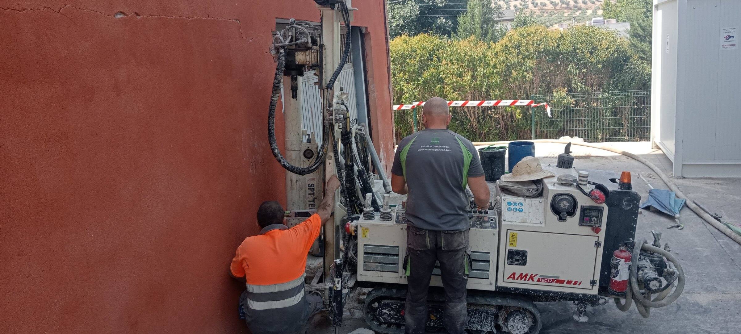 Dos operarios junto a una perforadora compacta sobre orugas trabajando al pie de la fachada del edificio.