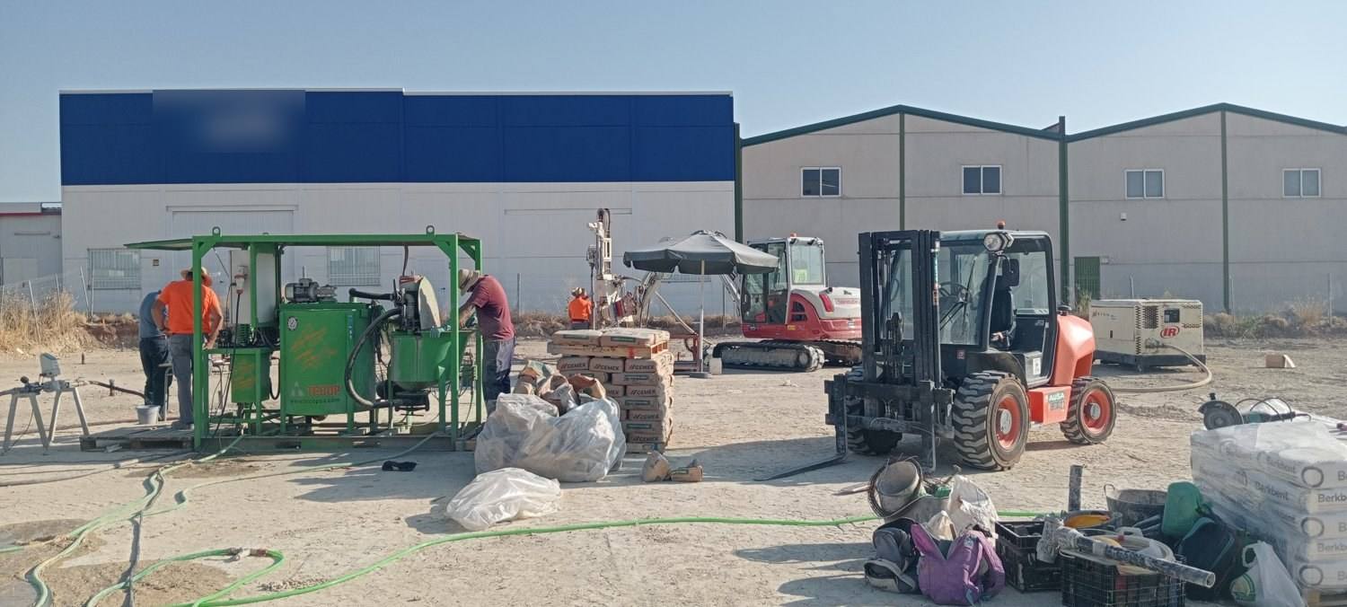 Central de inyección verde con operarios, palés de cemento, una perforadora sobre cadenas, una carretilla elevadora y un compresor en el solar.