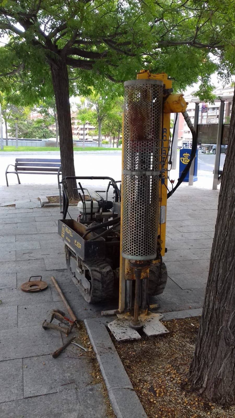 Penetrómetro dinámico sobre orugas en la acera junto a la glorieta, con el mástil de golpeo vertical entre dos árboles.