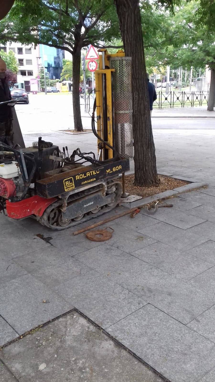 Penetrómetro dinámico sobre orugas en un alcorque de la acera, con el tráfico de la glorieta al fondo.