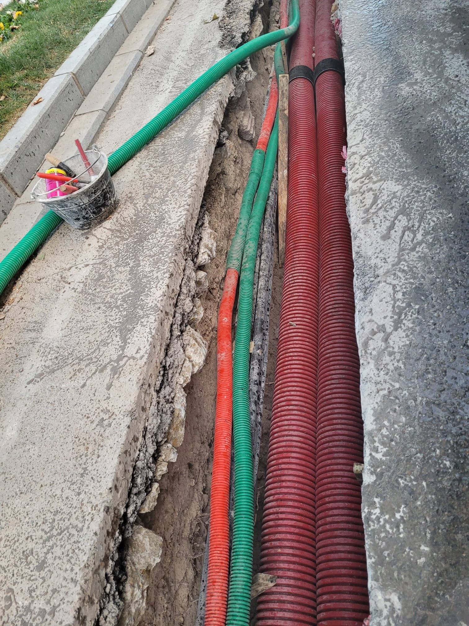 Zanja abierta junto a un bordillo con cuatro tubos corrugados rojos para cables y tubos verdes de telecomunicaciones a lo largo.