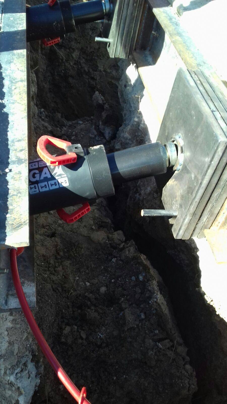 Cilindro hidráulico con manguera roja colocado en la zanja entre la viga de reacción y una placa de reparto apoyada en la zapata.