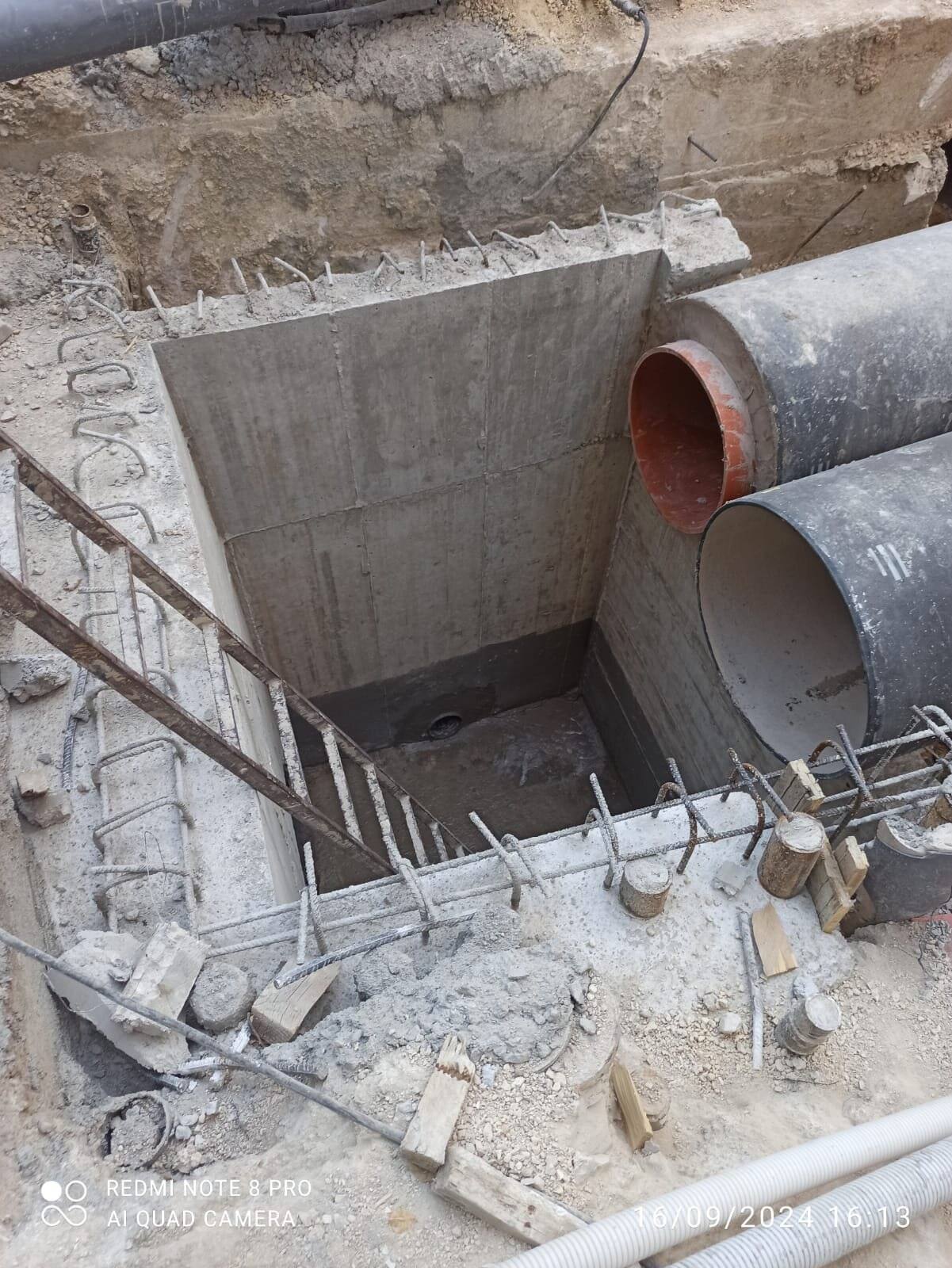 Arqueta de hormigón en ejecución con encofrado de madera y armadura, atravesada por dos tuberías.