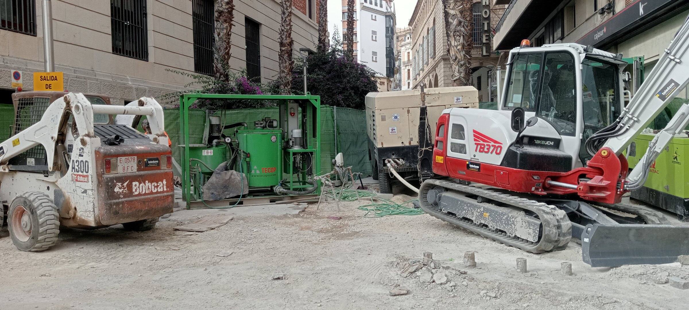 Calle del centro de Jaén vallada con malla verde, con una minicargadora, una excavadora de cadenas y equipos de obra entre edificios.