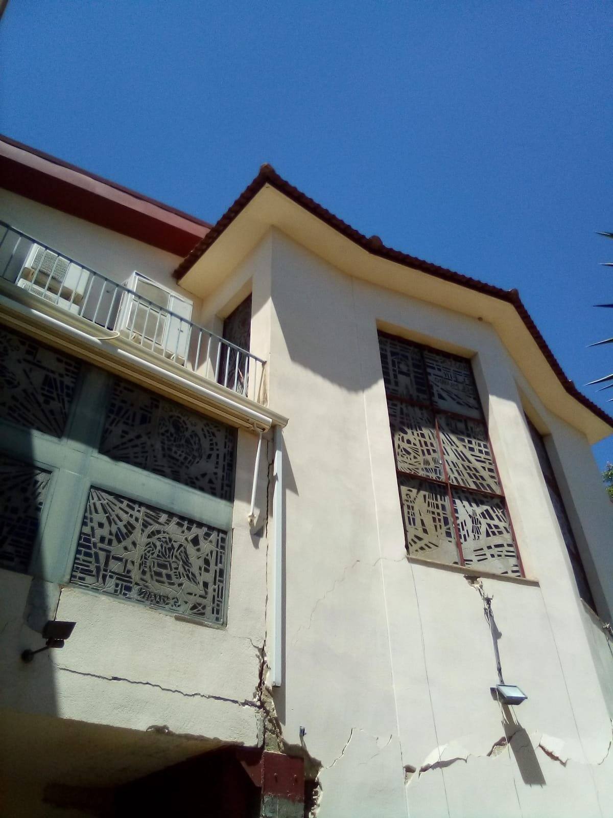 Esquina de la casa parroquial vista desde abajo, con balcón, celosías en las ventanas y una grieta en el zócalo de la fachada.