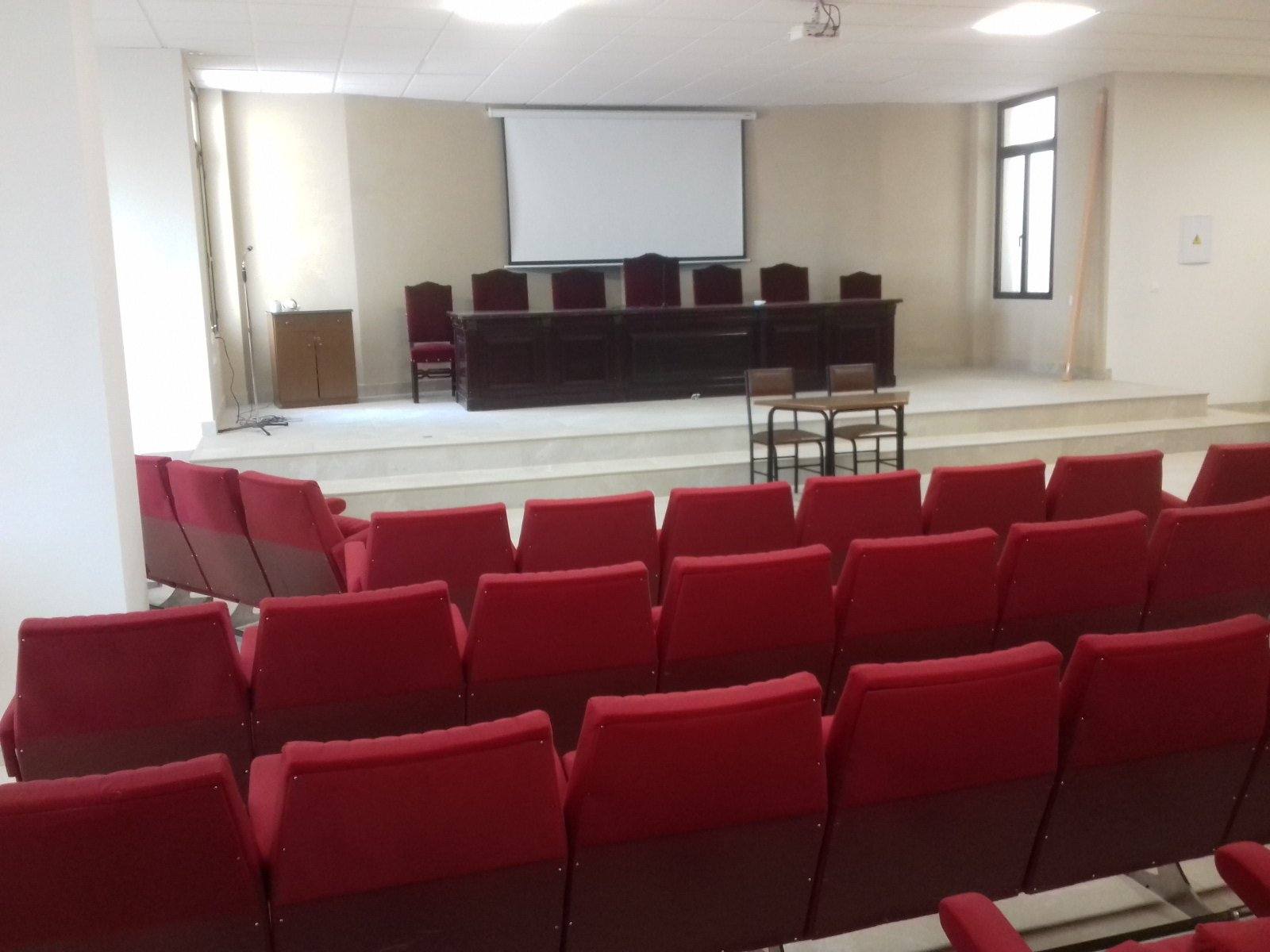 Salón de actos de la planta baja terminado, con filas de butacas rojas frente a una mesa y una pantalla.