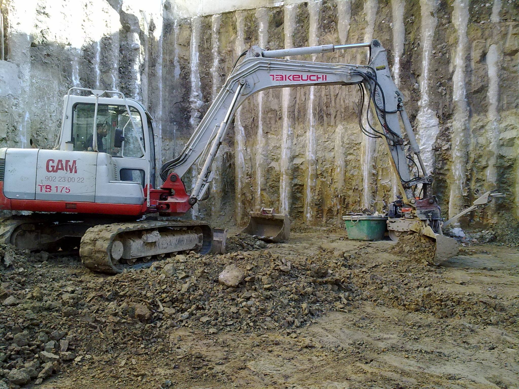 Excavadora de cadenas trabajando al pie de una pantalla de pilotes, con el terreno natural visible entre los pilotes.