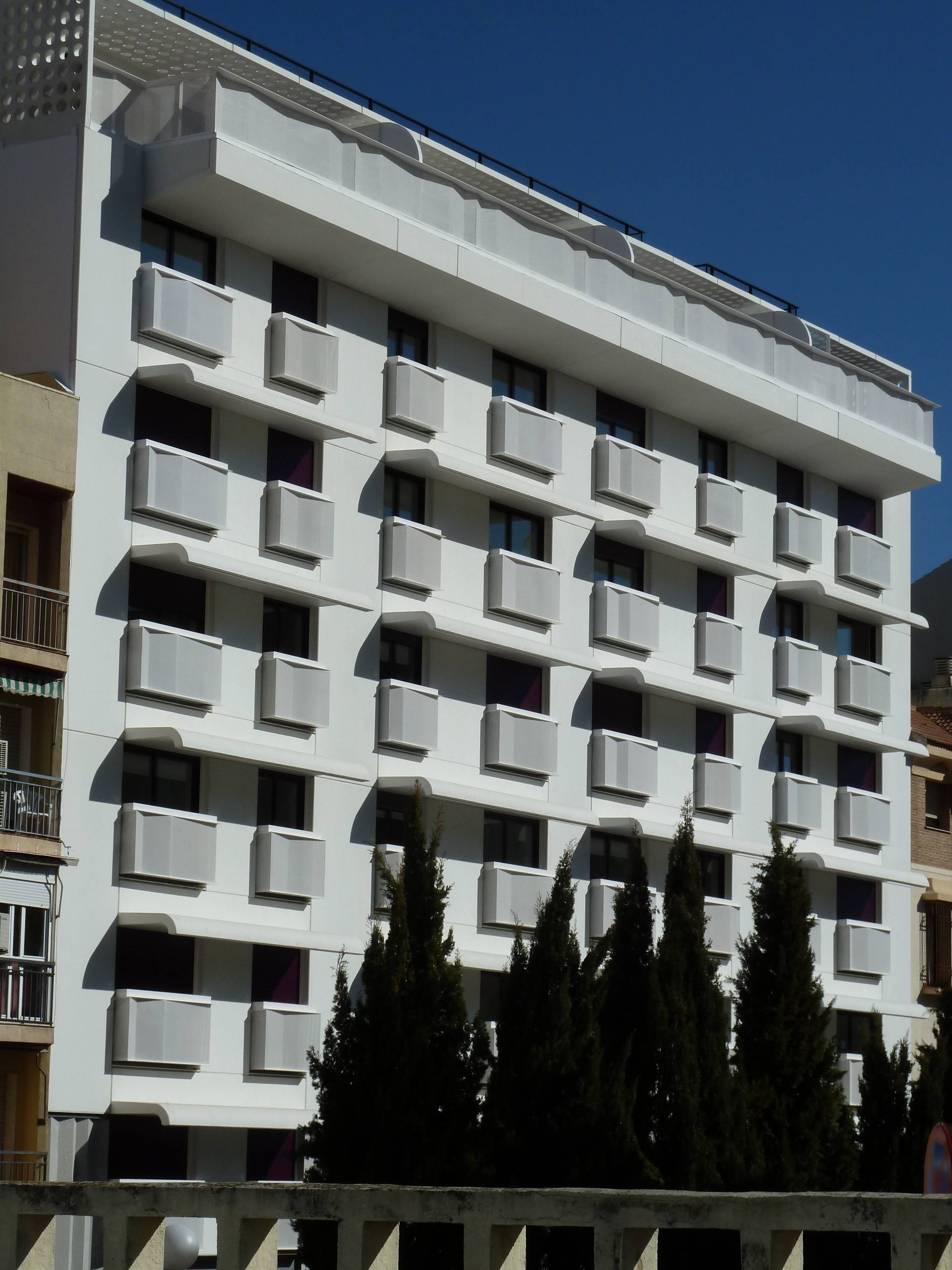 Edificio residencial de ocho plantas con terrazas continuas y antepechos claros, fotografiado contra un cielo azul.