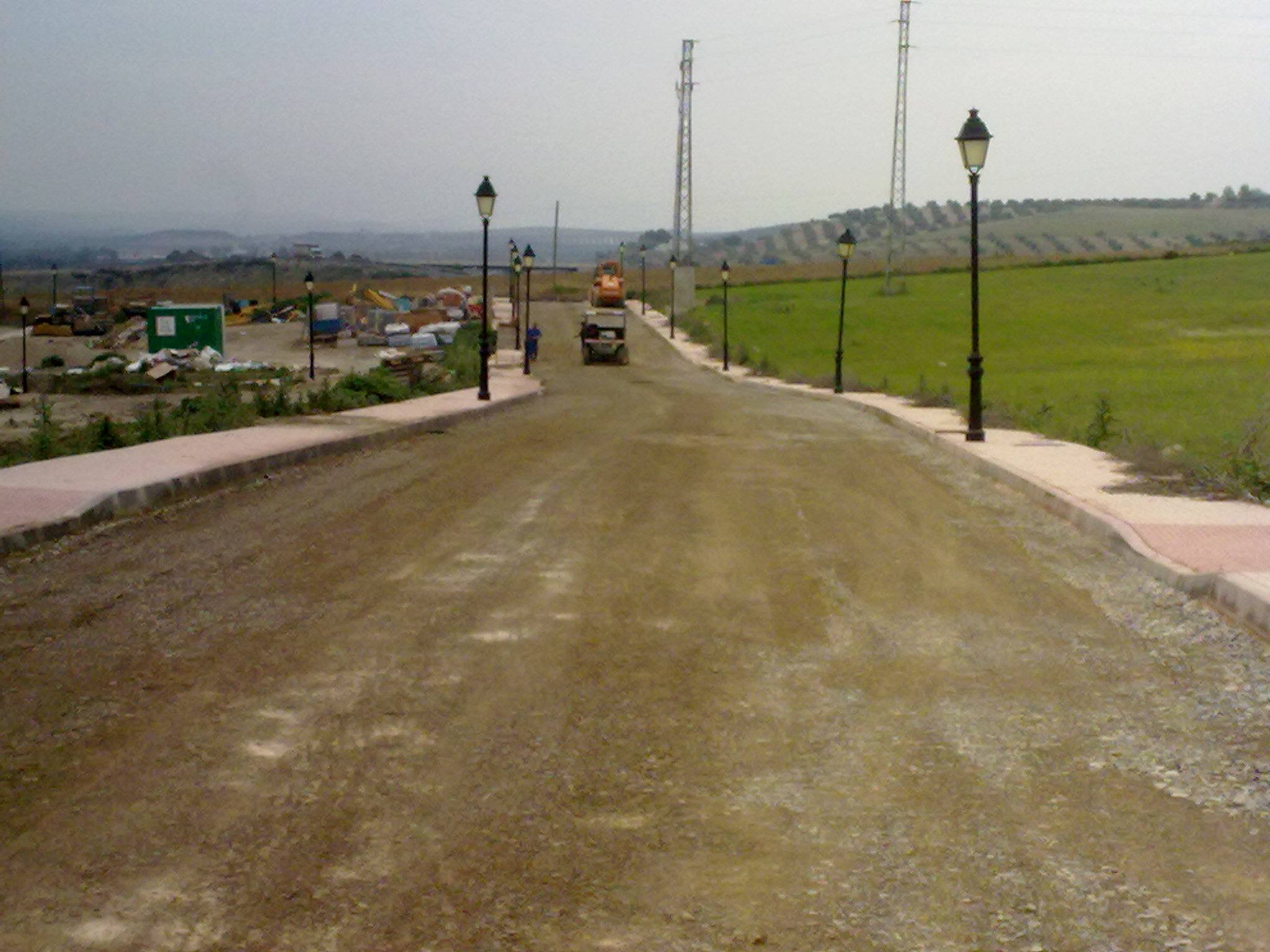 Vial de urbanización en ejecución con la explanada terminada, farolas instaladas y acera en construcción.