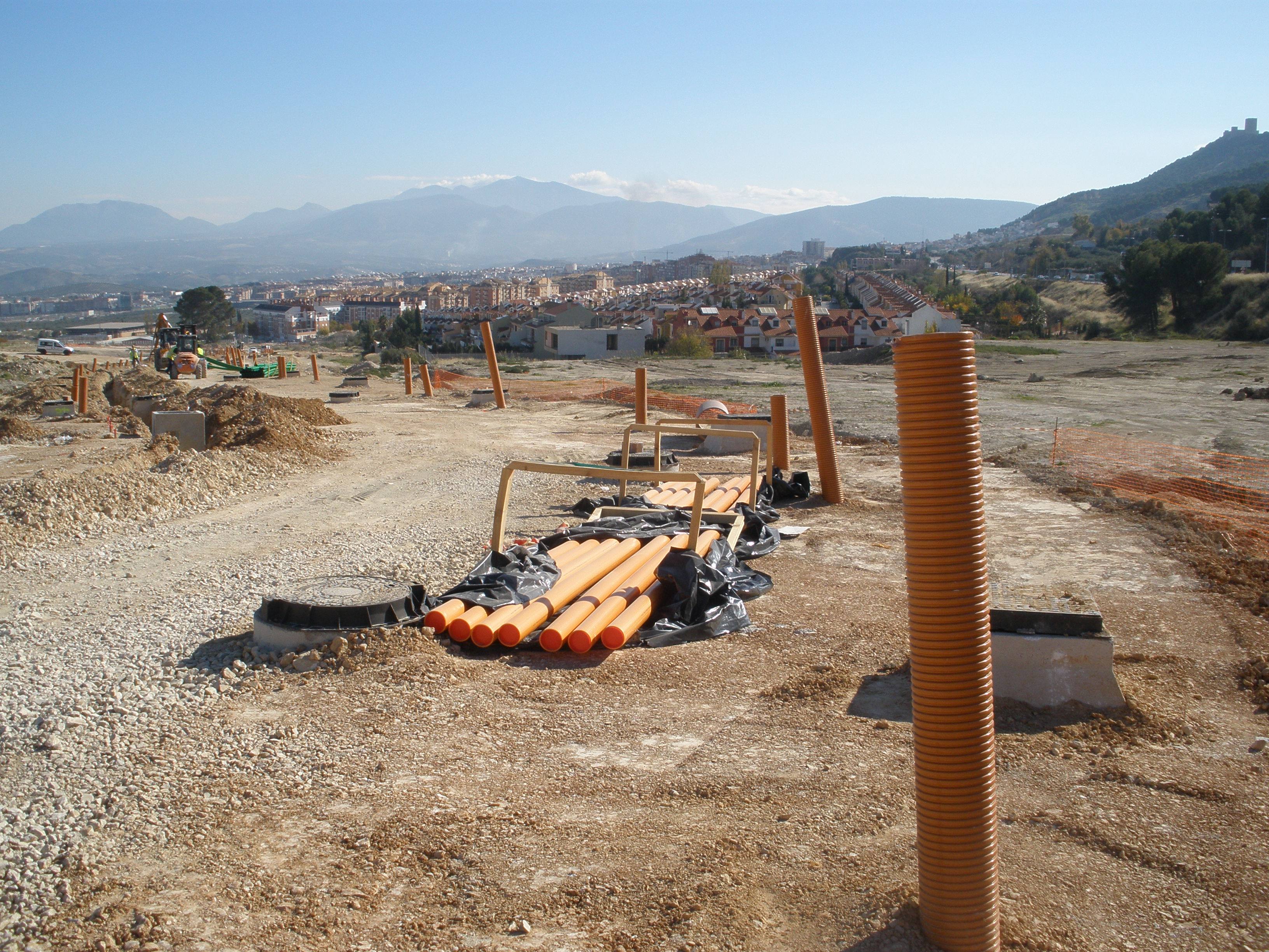 Obra de urbanización con tubos corrugados de saneamiento acopiados, arquetas y chimeneas de pozo en ejecución; al fondo, Jaén y Sierra Mágina.